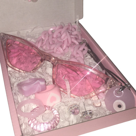 A premium pink boutique gift box filled with white crinkle paper shredded packaging, displaying a pair of translucent bubblegum pink cat-eye fashion glasses with a chunky pink chain lanyard strap, a set of pink and lavender marbled acrylic rings including a heart design, pink rhinestone heart stud earrings, and a matching pink evil eye charm pendant.