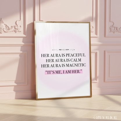 A framed typographic art print has a white background with a soft pink glow and black text reading, "HER AURA IS PEACFUL HER AURA IS CALM HER AURA IS MAGNETIC 'IT'S ME, I AM HER.