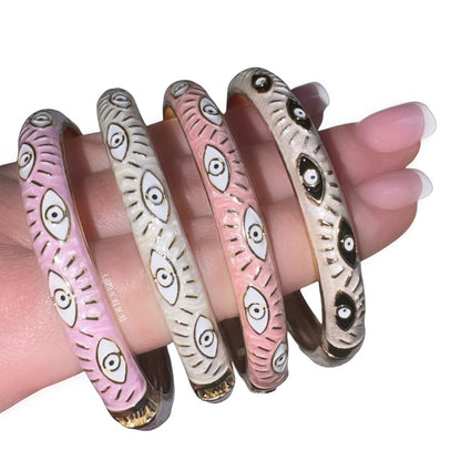 Close-up view of a hand with a classic french manicure showcasing a stacked collection of four enamel evil eye bangles in contrasting pastel pink and creamy white shades, detailed with gold accents and repeating protective eye motifs.