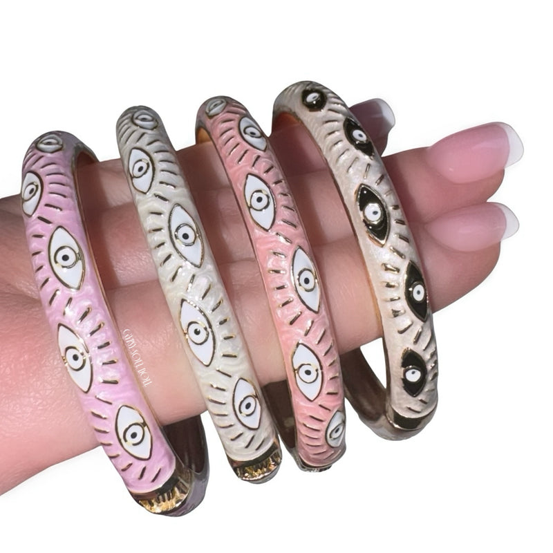 Close-up view of a hand with a classic french manicure showcasing a stacked collection of four enamel evil eye bangles in contrasting pastel pink and creamy white shades, detailed with gold accents and repeating protective eye motifs.