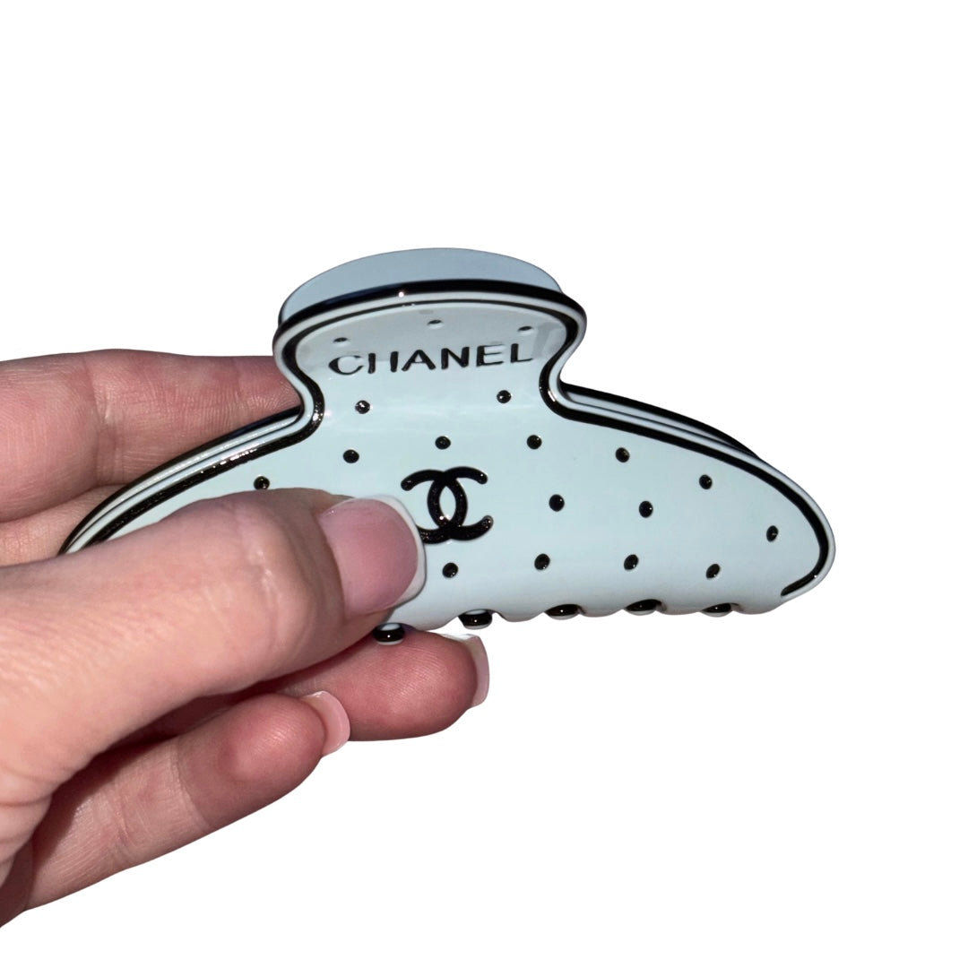 A hand holding a blue Chanel hair claw clip with black trim, black polka dots, and the Chanel logo and wordmark on the front, against a plain white background.