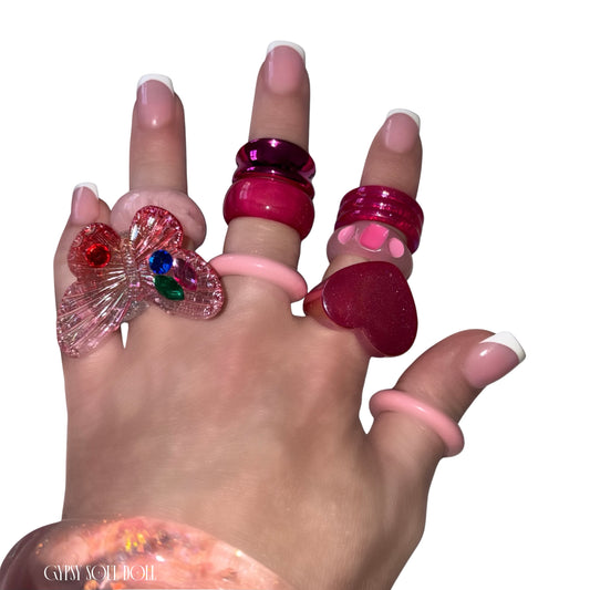 A close-up view of a hand with French tip manicures crossing over each other, modelling a chunky pink and red acrylic candy ring stack by Gypsy Soul Doll that features a large translucent pink 3D butterfly statement ring, a glossy red heart ring, and stacked pastel pink resin bands against a clean white background.