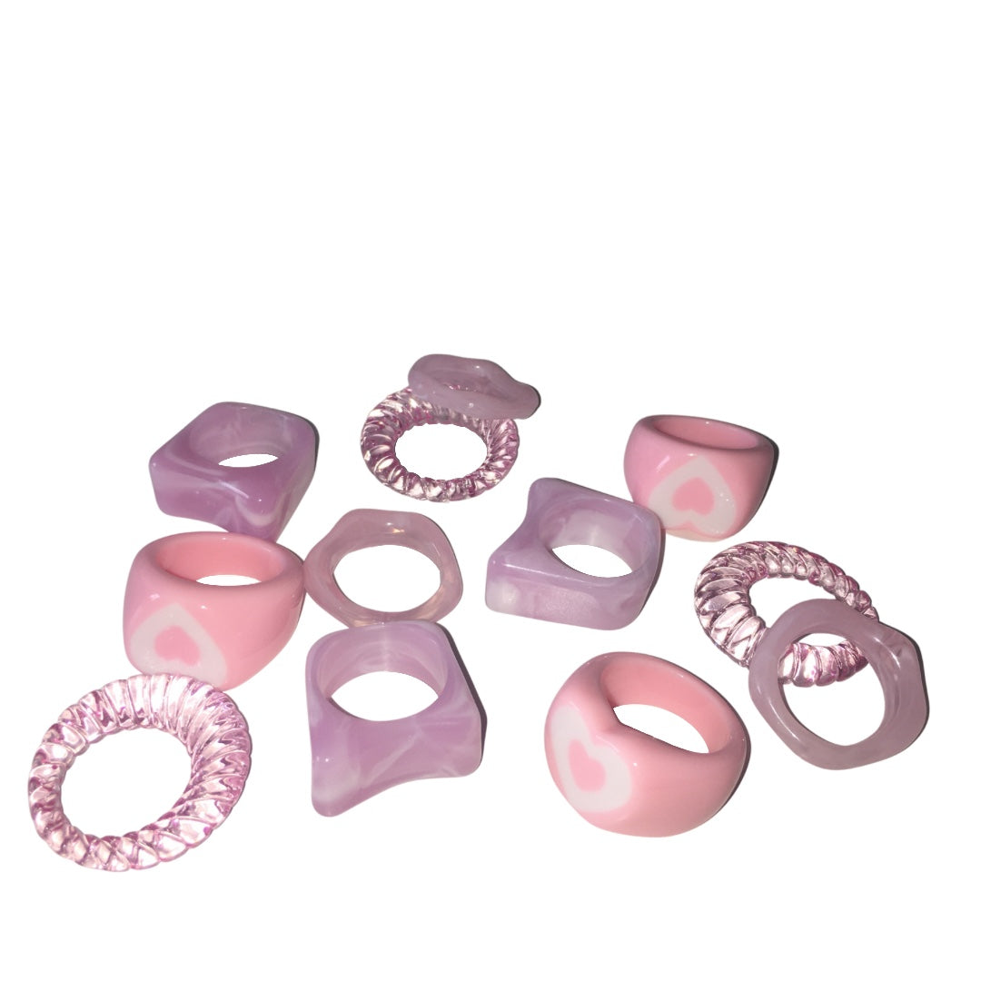 A collection of eleven chunky acrylic resin rings scattered on a clean white background, showcasing a mix of pastel pink heart dome rings, marbled lavender square block rings, clear pink twisted spiral bands, and wavy translucent purple jelly rings.