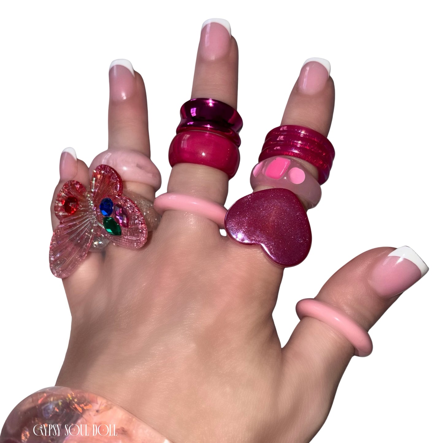 A close-up view of a hand with French tip manicures crossing over each other, modelling a chunky pink and red acrylic candy ring stack by Gypsy Soul Doll that features a large translucent pink 3D butterfly statement ring, a glossy red heart ring, and stacked pastel pink resin bands against a clean white background.