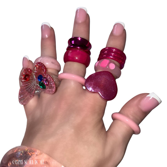 A close-up view of a hand with French tip manicures crossing over each other, modelling a chunky pink and red acrylic candy ring stack by Gypsy Soul Doll that features a large translucent pink 3D butterfly statement ring, a glossy red heart ring, and stacked pastel pink resin bands against a clean white background.