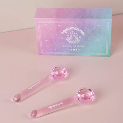 Two pink cooling ice globes with clear stems and spherical ends, displayed in front of a pastel iridescent box labeled aquamarina on a soft pink surface.