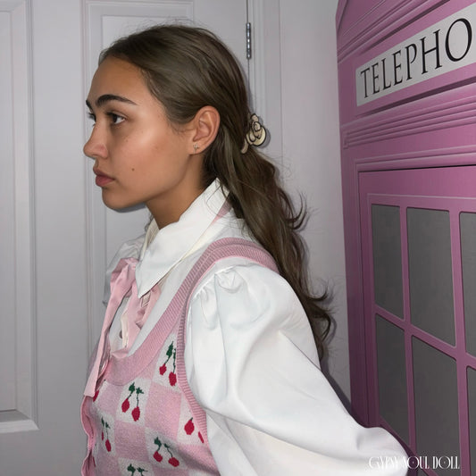 A side profile view of a model standing next to a bright pink telephone booth, showcasing her wavy brown hair styled in a half-up look secured with a metallic shell-shaped hair claw clip, paired with a cherry knit vest by Gypsy Soul Doll.