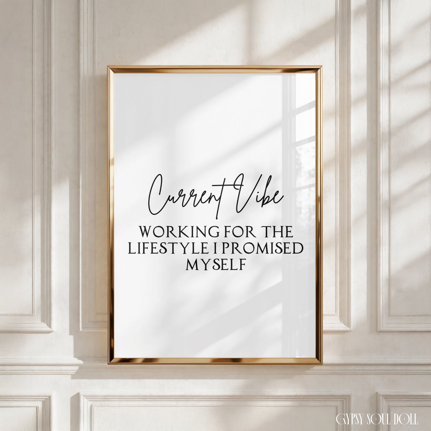 A framed typographic wall art print with a gold frame hangs on a white paneled wall. The print has a white background with black text reading 'Current Vibe' and 'WORKING FOR THE LIFESTYLE I PROMISED MYSELF.