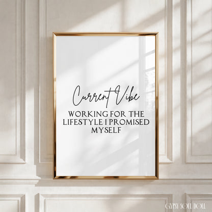 A framed typographic wall art print with a gold frame hangs on a white paneled wall. The print has a white background with black text reading 'Current Vibe' and 'WORKING FOR THE LIFESTYLE I PROMISED MYSELF.