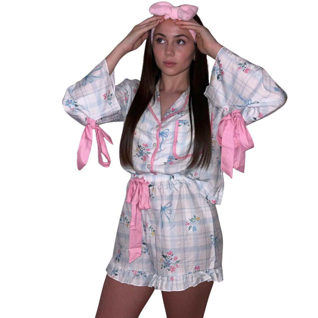 A portrait view of a model with straight brunette hair adjusting a plush pink bow headband against a plain white background, showcasing a luxury light blue and white checked satin pyjama shorts set featuring pastel floral details and large pink ribbon ties on the sleeves and waist by Gypsy Soul Doll.