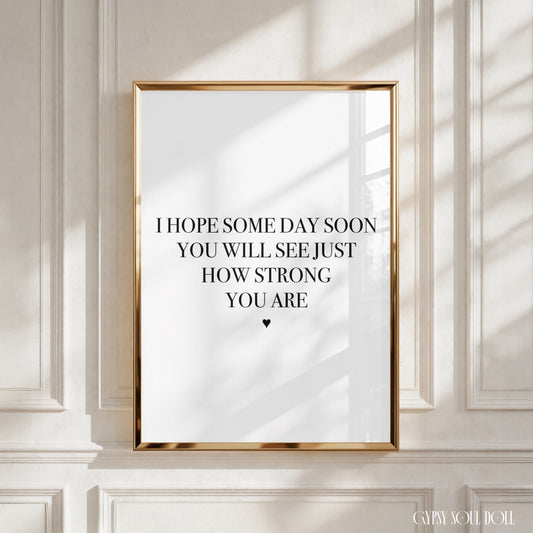 A framed motivational art print with the text 'I HOPE SOME DAY SOON YOU WILL SEE JUST HOW STRONG YOU ARE' and a small heart, displayed on a white paneled wall with soft window shadows.