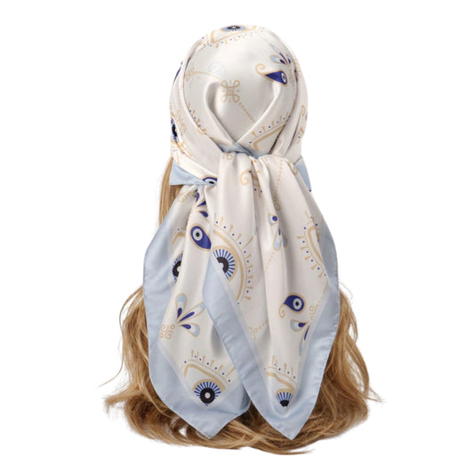Rear view of a model with long hair wearing an off-white silk-like satin square head scarf featuring a light blue border and a protective dark blue evil eye talisman pattern for an Ibiza festival style.