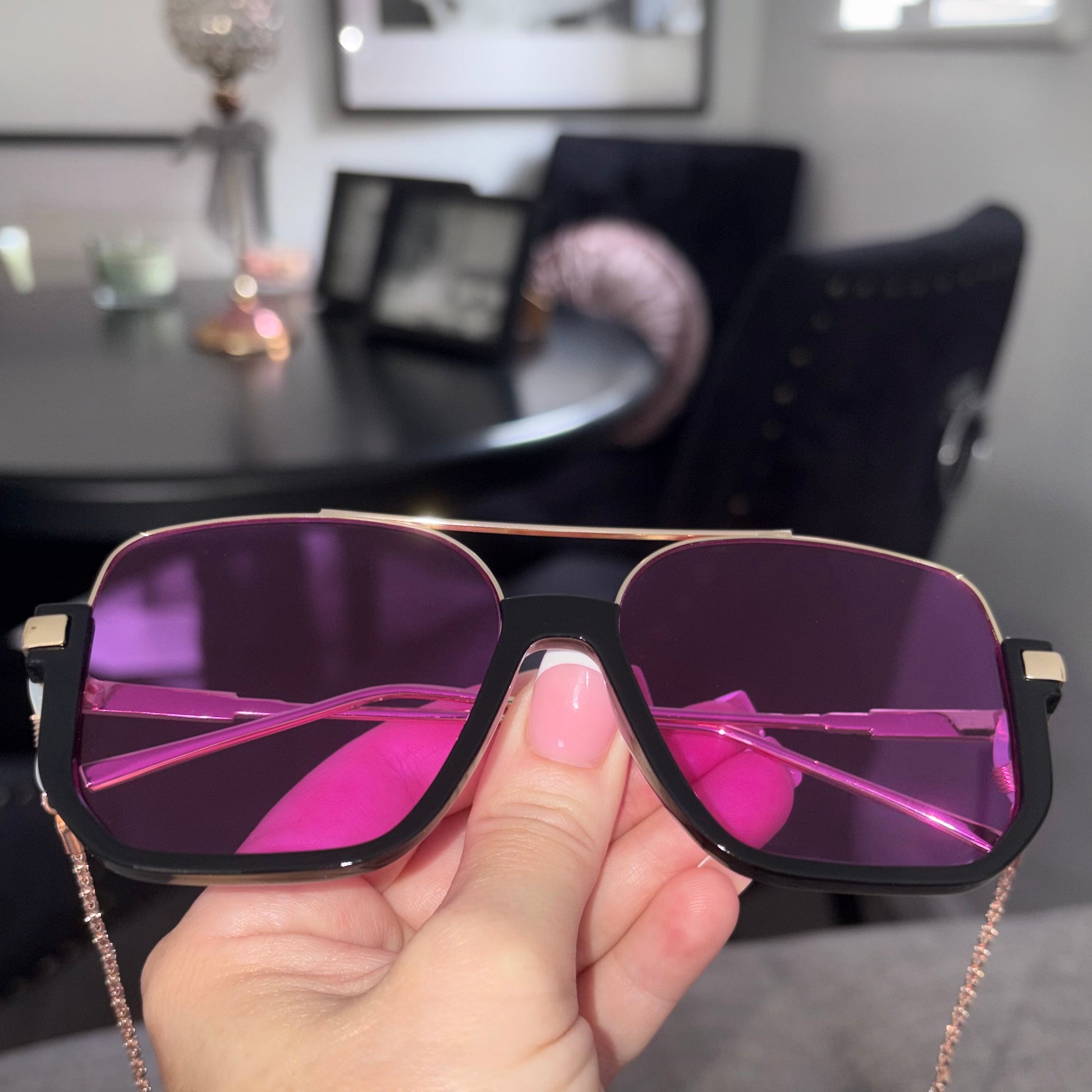 A hand holding a pair of oversized square sunglasses with a black frame, rose gold metal trim, and vibrant purple mirrored lenses, set against a blurred dining room background.