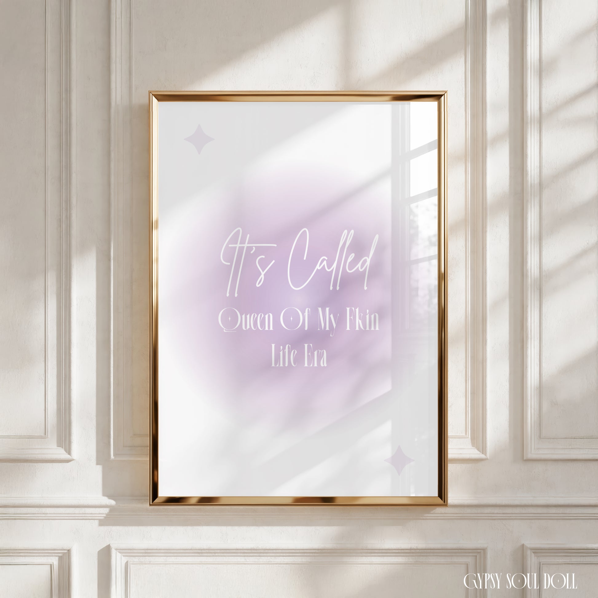A framed art print with a gold frame hangs on a white paneled wall. The print features a soft lavender and white watercolor background with the text 'It's Called Queen Of My Own Life Era' in white script and serif fonts.
