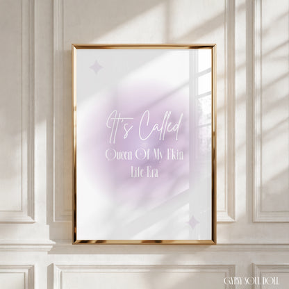 A framed art print with a gold frame hangs on a white paneled wall. The print features a soft lavender and white watercolor background with the text 'It's Called Queen Of My Own Life Era' in white script and serif fonts.