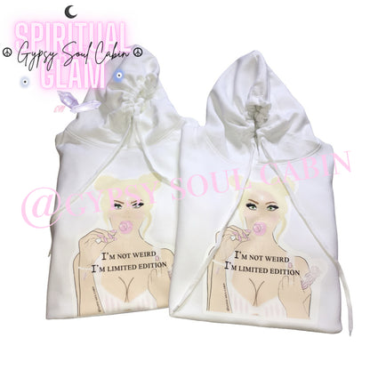 Two white oversized hoodies with a graphic of a woman with blonde hair, holding a lollipop, and the text "I'M NOT WEIRD, I'M LIMITED EDITION" printed on the front. The design features pastel colours and a playful, artistic style.