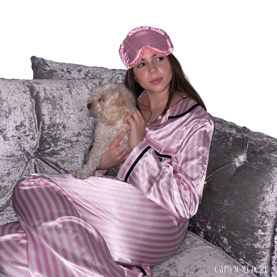 A model lounging on a grey crushed velvet sofa while holding a small fluffy white dog, dressed in a pink and white candy stripe luxury satin pyjama set with a matching ruffled pink satin sleep mask on her head against a white background by Gypsy Soul Doll.