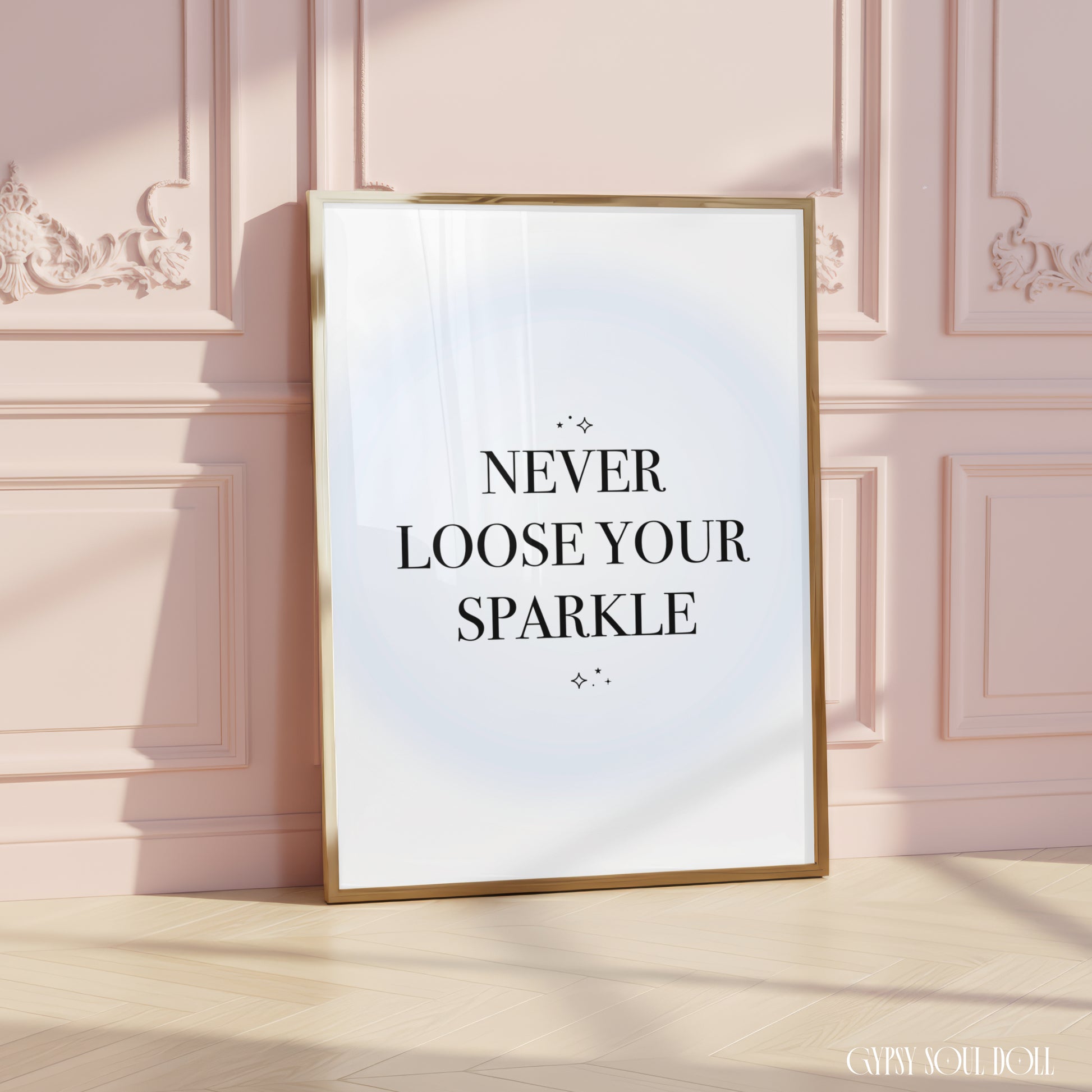 A framed typographic wall art print leaning against a pale pink paneled wall on a light wood floor. The print has black serif text reading “NEVER LOOSE YOUR SPARKLE” on a white background with a soft pale blue glow effect, in a thin gold frame.