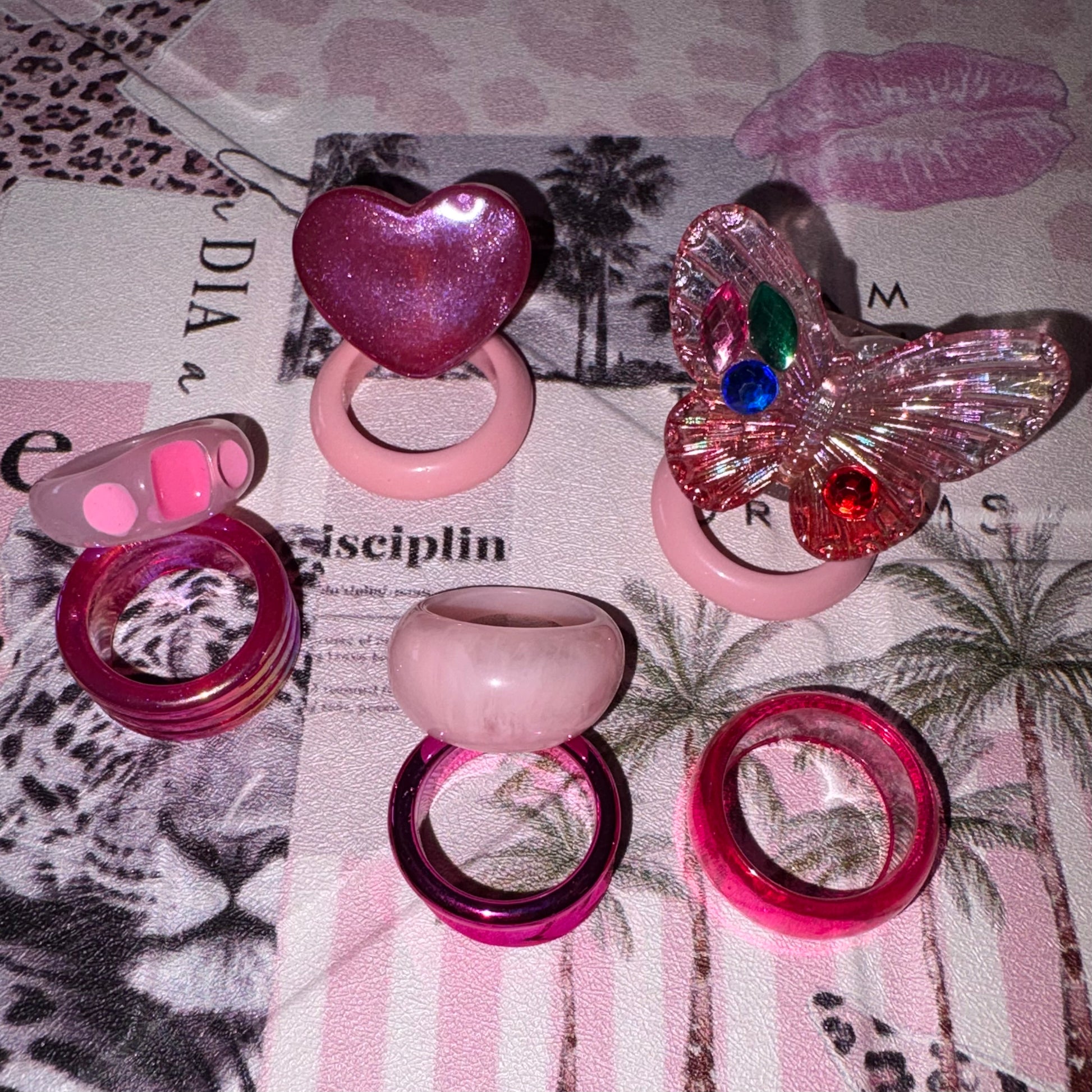 A set of pink-themed novelty rings. The set includes a glittery pink heart ring, a butterfly-shaped ring with rhinestones, a translucent pink ring, a glossy pink ring for Ibiza or Festivals.