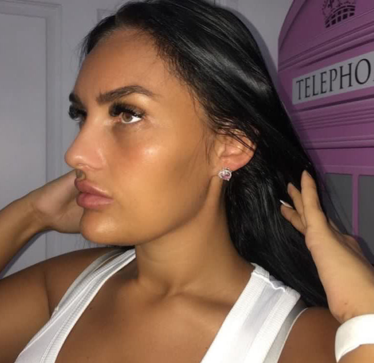 Side profile view of a woman with long dark hair modelling the Gypsy Soul Doll pink rhinestone heart stud earrings, showcasing the sparkling silver-toned crystal halo design against a backdrop featuring a pink telephone box.