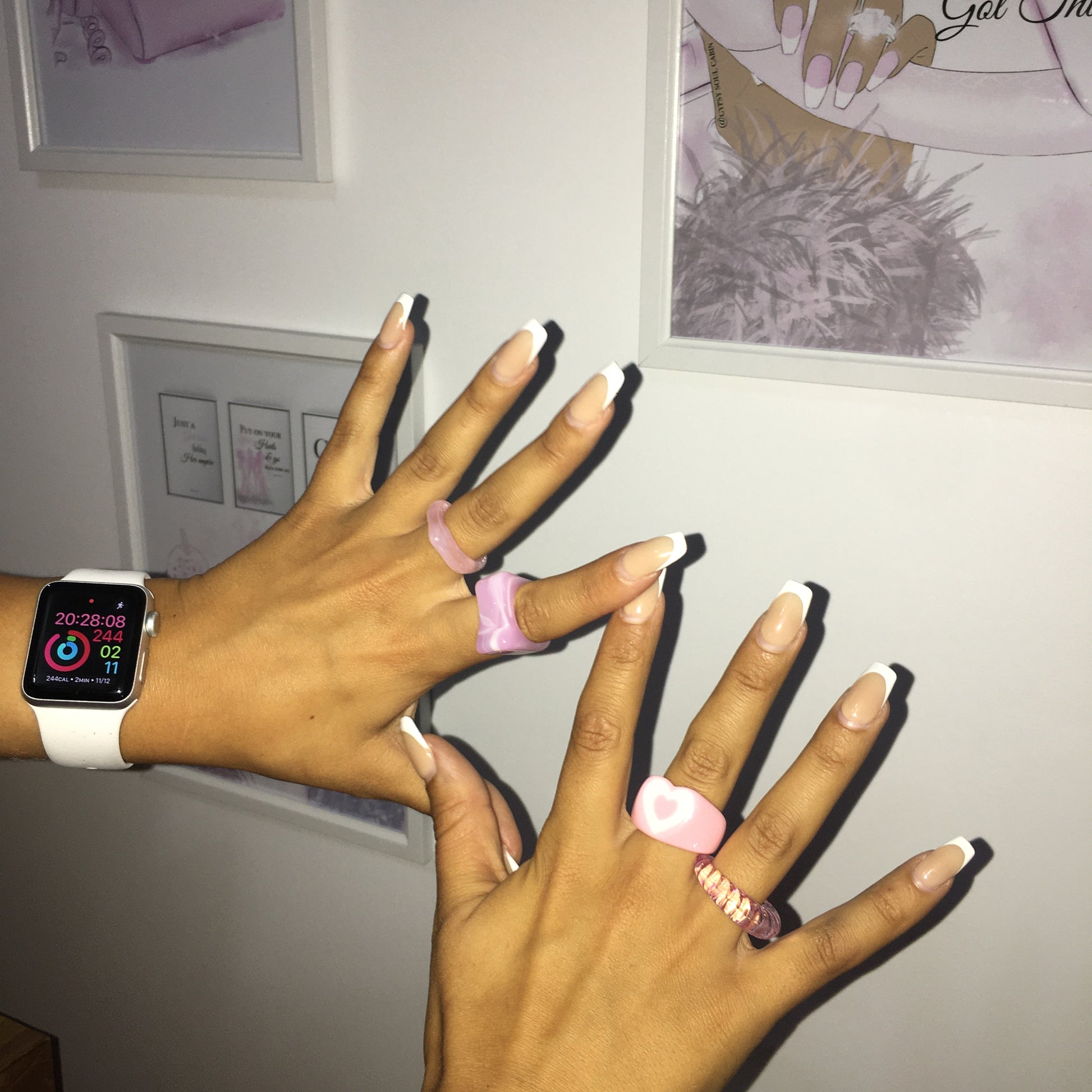 A close-up view of two hands with long French tip manicures crossed over each other, modeling the GSD chunky resin heart ring set featuring a pastel pink heart dome ring, a clear pink twisted textured spiral band, and marbled lavender acrylic block rings against a background of framed pink wall art by Gypsy Soul Doll.