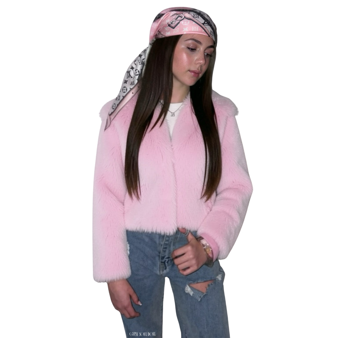 A woman modelling a light pink faux fur jacket, styled with a pink and black patterned monogram logo bandana, a white t-shirt, and distressed blue jeans against a plain white background.
