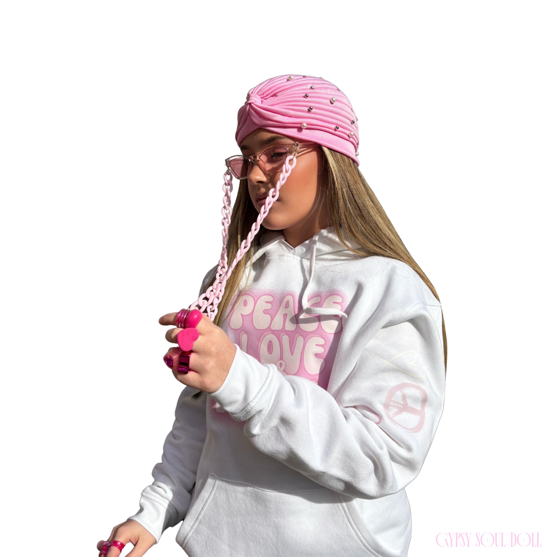A model wearing a pink boho head wrap with pink fashion glasses, a chain strap attached, a white hoodie with pink text, and pink chunky retro rings against a plain white background.
