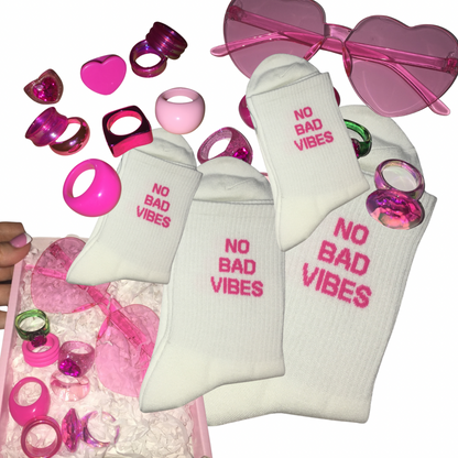 White crew socks with pink 'NO BAD VIBES' text, displayed with pink heart-shaped fashion glasses, pink and green ring-shaped accessories, and a pink gift box on a white background.