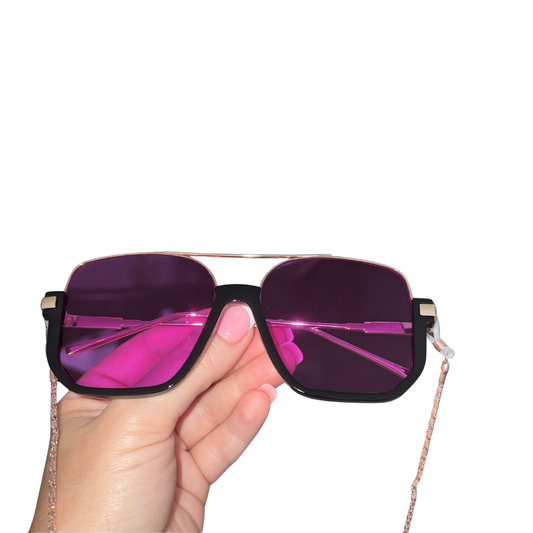 A hand holding a pair of oversized square sunglasses with a black frame, rose gold double bridge, and vibrant purple mirrored lenses against a solid white background.