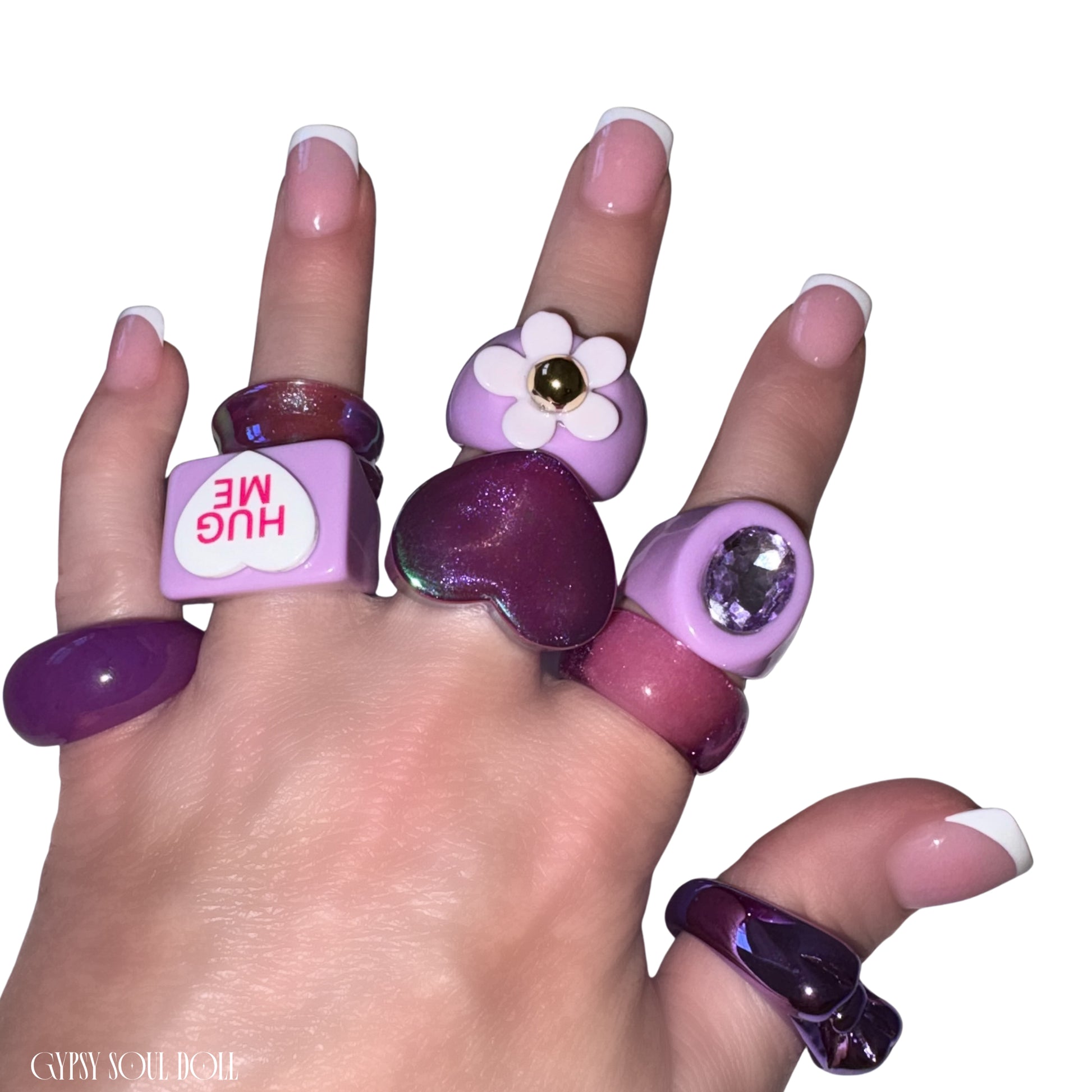 A close-up view of a hand with French-tip manicures crossing over each other, modelling a chunky pastel purple acrylic candy ring stack by Gypsy Soul Doll that features a matte lilac 3D flower ring, a square 'HUG ME' conversation heart ring, a glossy translucent plum heart ring, and chunky resin bands with embedded violet rhinestones against a clean white background.