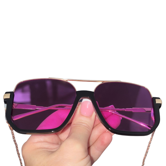A hand holding a pair of oversized square sunglasses with dark purple lenses, a black frame, rose gold metal double bridge and temples, and a thin rose gold chain strap, shown against a plain white background.