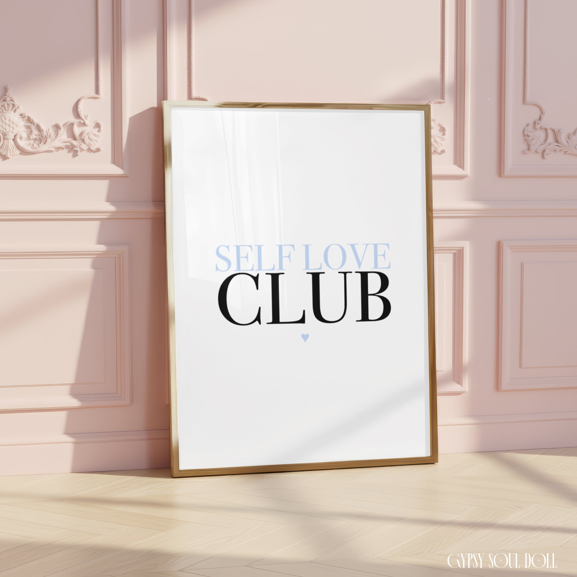 A framed typographic art print leaning against a pale pink paneled wall on a light wood floor. The print has a white background with the words 'SELF LOVE CLUB' in black and light blue, and a thin gold frame.