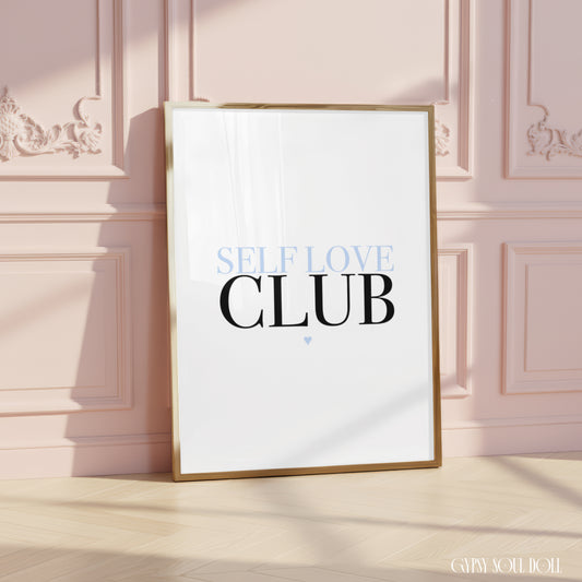 A framed typographic art print leaning against a pale pink paneled wall on a light wood floor. The print has a white background with the words 'SELF LOVE CLUB' in black and light blue, and a thin gold frame.