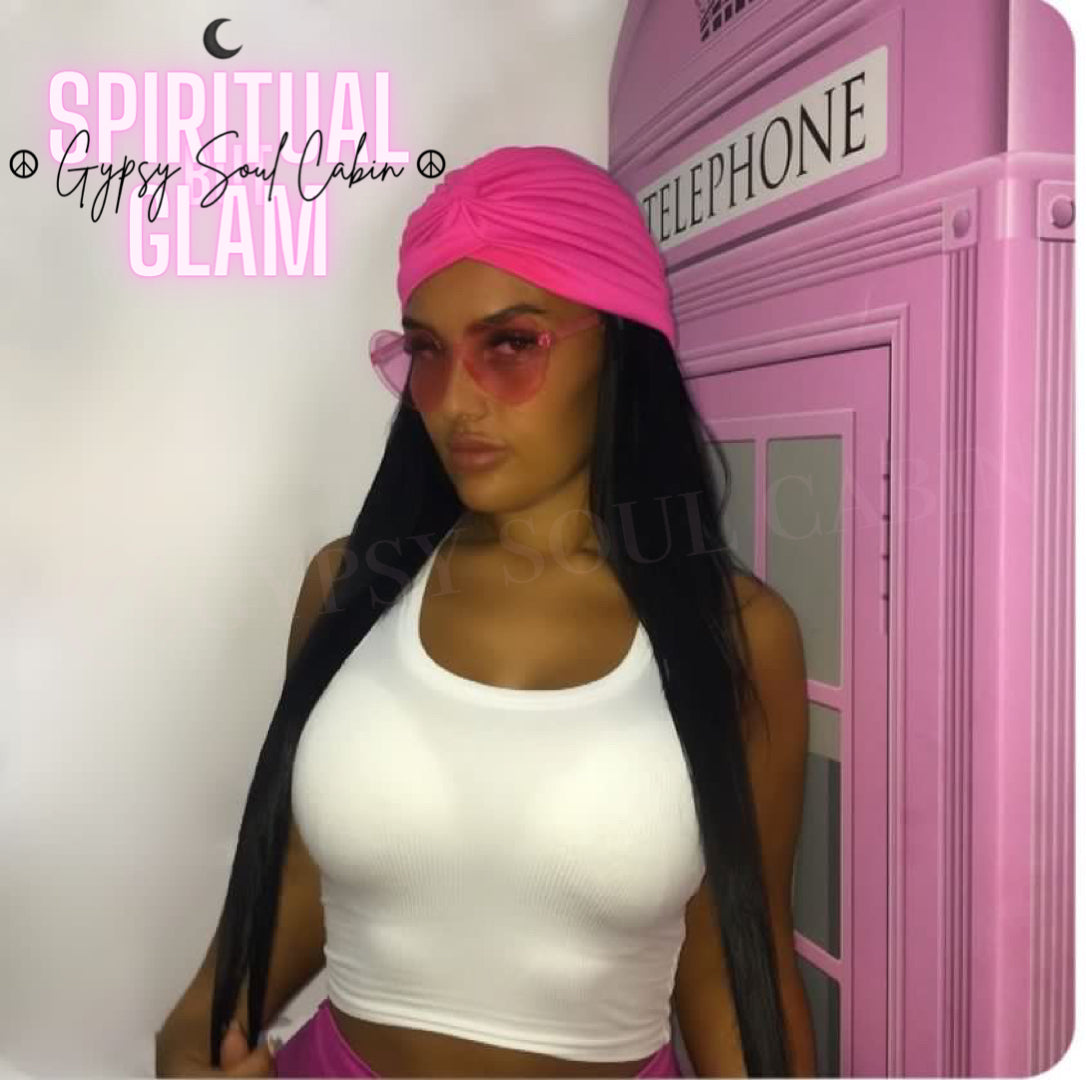 A woman wearing a pink Bohemian gypsy head wrap with a white top, pink trousers and fashion Y2K glasses standing in front of a pink telephone booth.