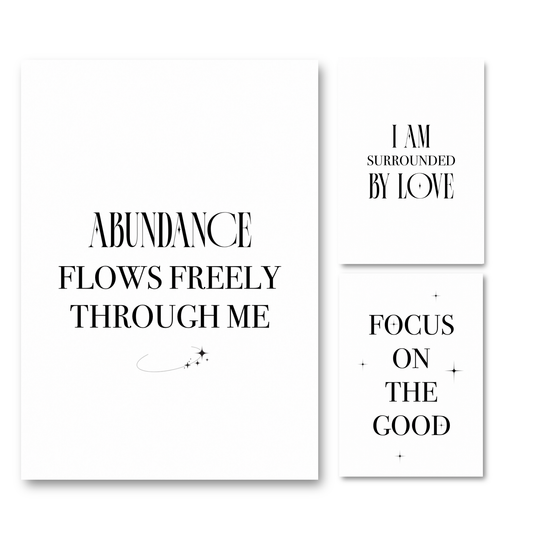 Three minimalist white art prints with black typography on a white background. The largest print reads 'ABUNDANCE FLOWS FREELY THROUGH ME' with a small curved line and star motif.
