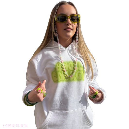 A woman modelling a white hoodie with a bright lime green 'ANGEL ENERGY' graphic on the chest, styled with neon green sunglasses, a chain lanyard, matching wrist accessories and chunky Y2K fashion rings against a plain white background.