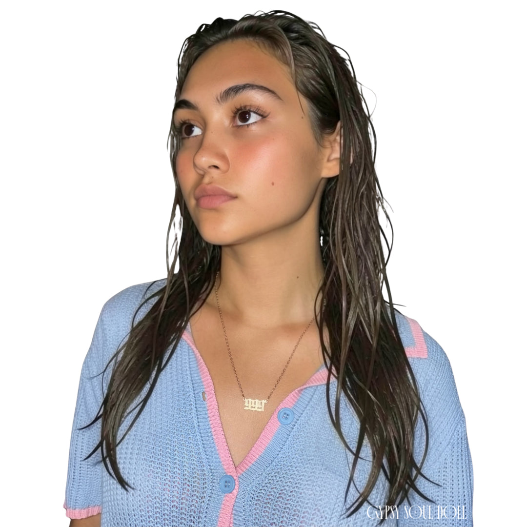 A model with wet-look hair wearing a blue and pink knit polo cardigan showcasing a gold 999 angel number nameplate necklace by Gypsy Soul Doll against a clean white backdrop.