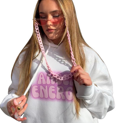 A young woman with long blonde hair wearing a white hoodie with a pink gradient 'ANGEL ENERGY' graphic and a chunky pink chain lanyard attached to Pink fashion glasses. She is also wearing 6 retro chunky rings posing against a solid white background.