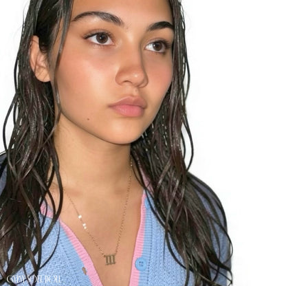 A close-up of a woman with wet, dark hair wearing a light blue-and-pink knit cardigan and a delicate gold necklace with an angel number 111 pendant, set against a plain white background.
