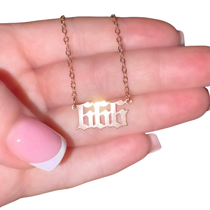 A close-up of a hand holding a delicate rose gold chain necklace with a nameplate pendant featuring the gothic-style angel number '666'.