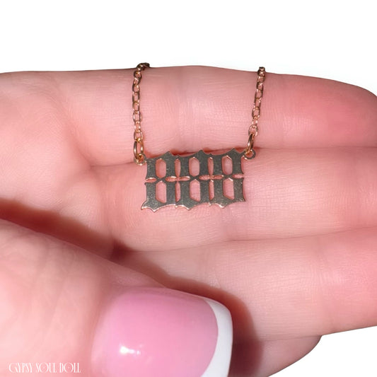 A close-up of a hand holding a delicate rose gold angel number 888 chain necklace with a gothic-style cutout pendant.
