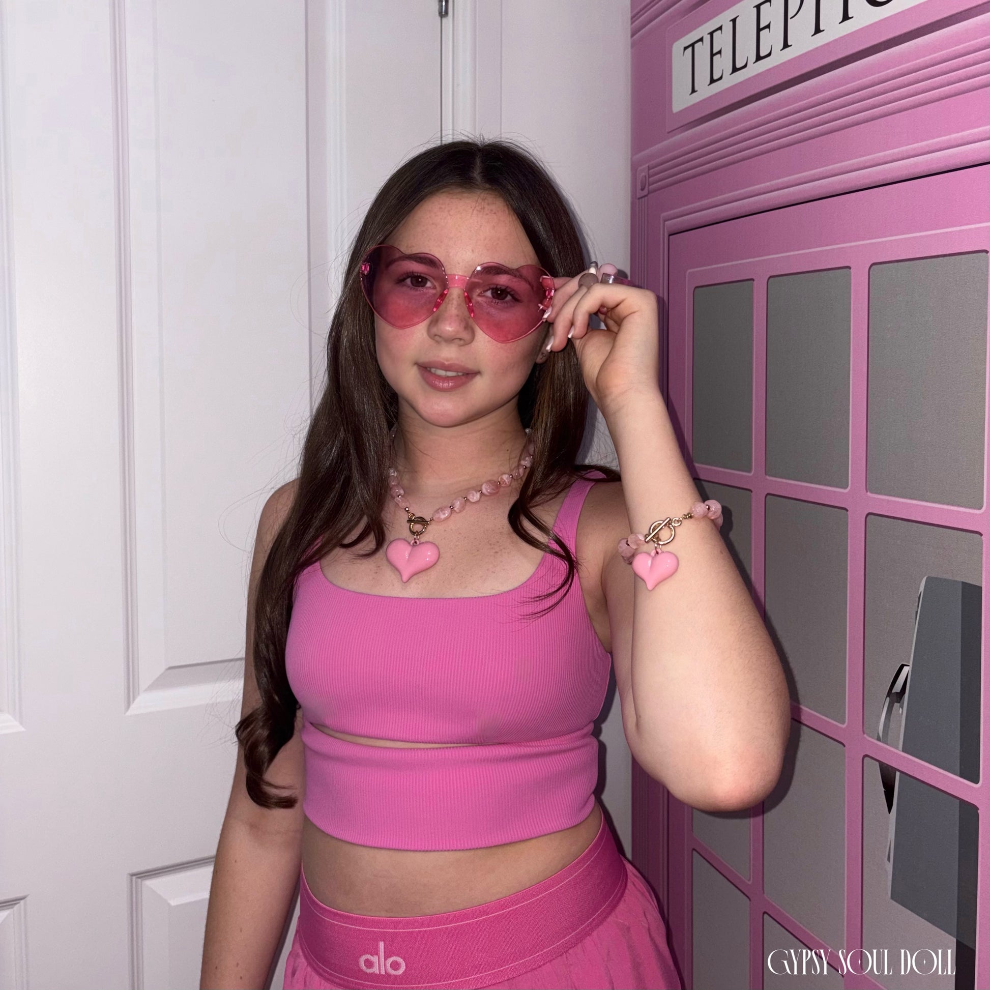 Model posing in front of a pink telephone box wearing the Gypsy Soul Doll Barbie World coordinates set, featuring pink heart sunglasses, a matching heart charm crystal necklace, and a bracelet stack.