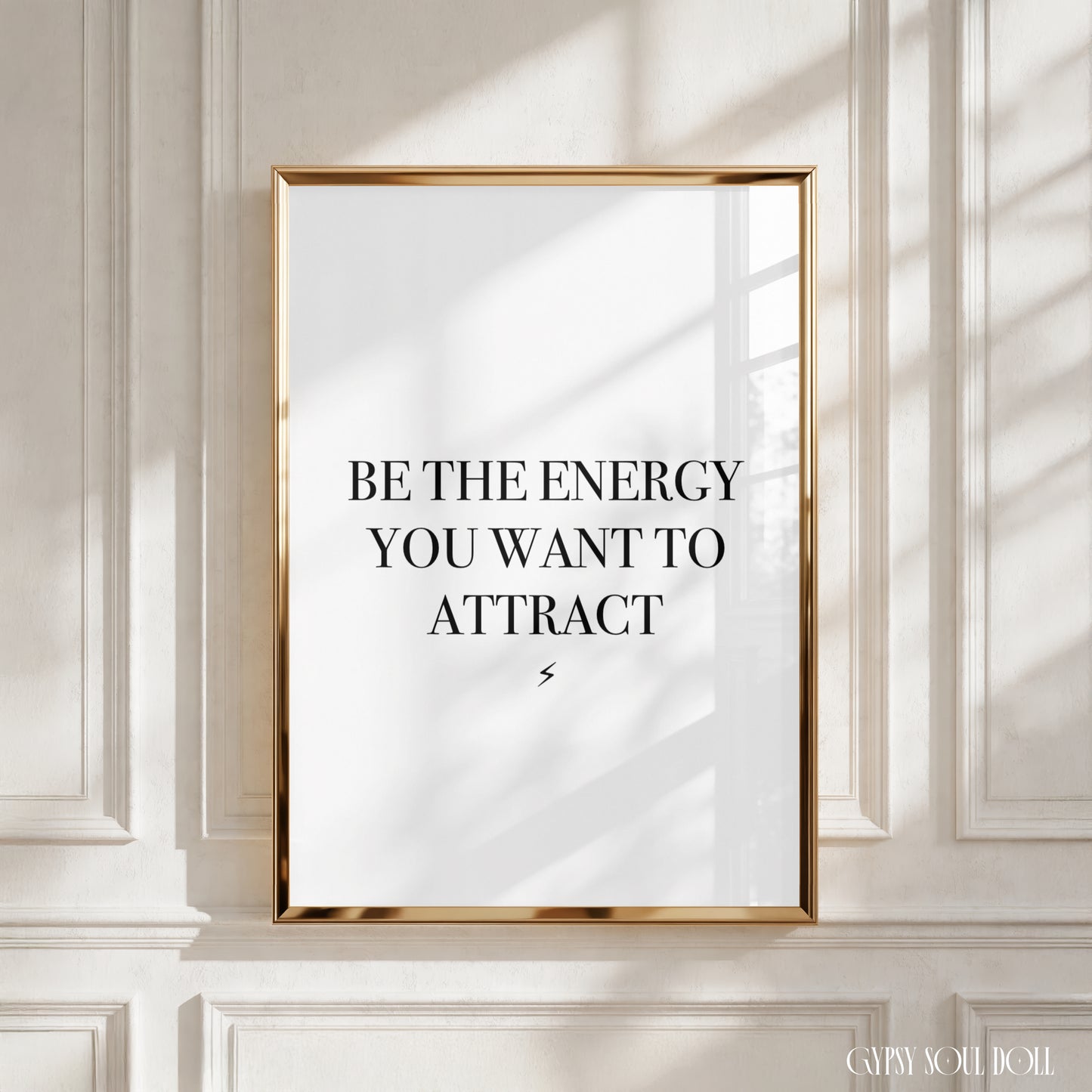 A framed typographic wall art print with the text 'BE THE ENERGY YOU WANT TO ATTRACT' in black serif lettering on a white background, set in a thin gold frame and displayed against a white paneled wall with soft window shadows.