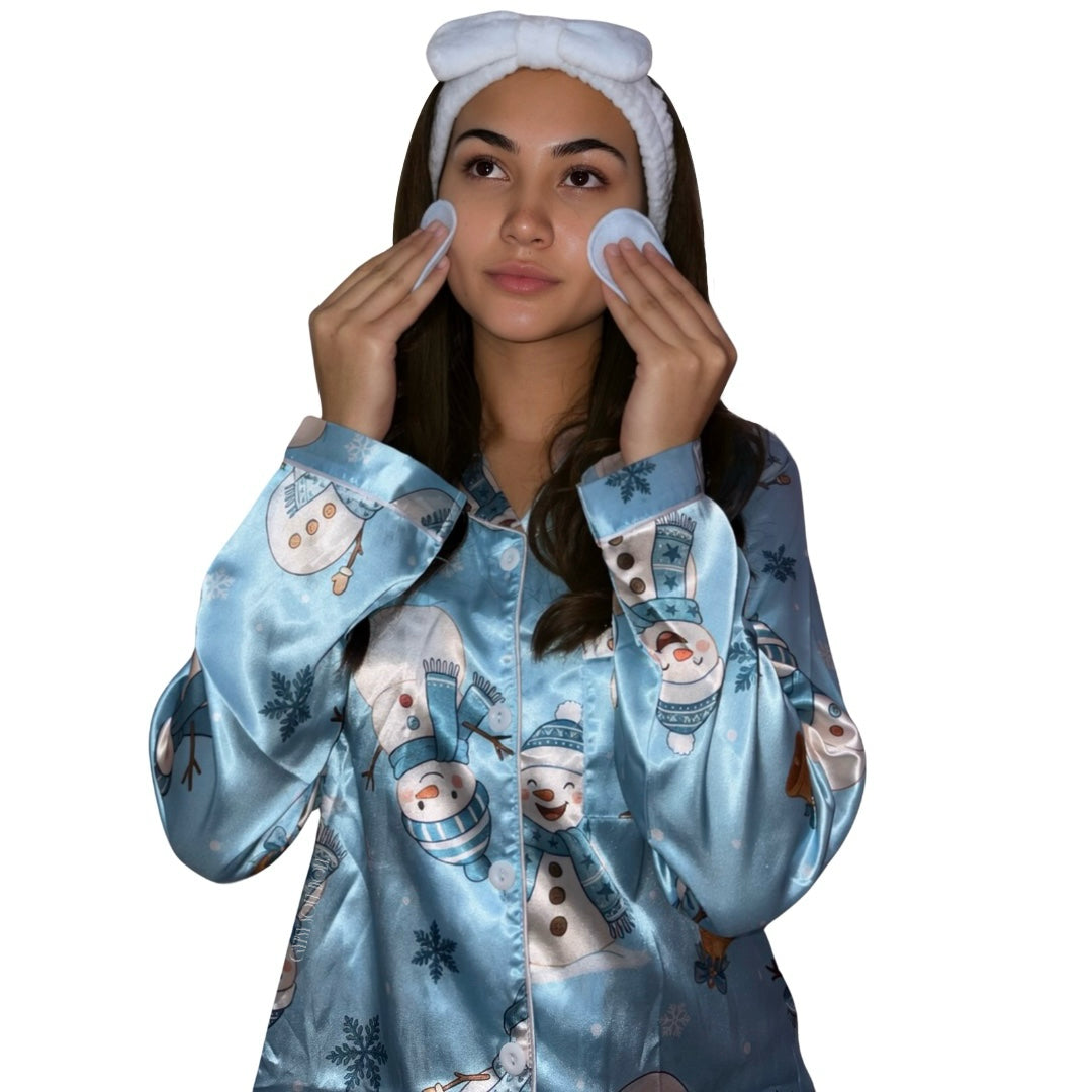 A woman wearing a light blue satin pyjama set with a snowman print and a matching white headband, holding blue towel facial pads near her face against a plain white background.