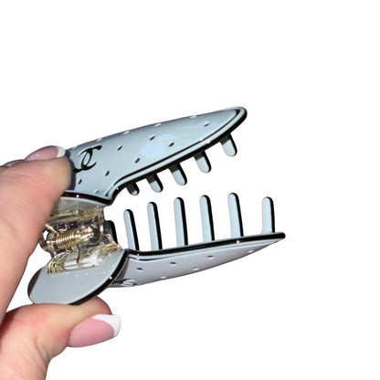 A blue Chanel hair claw clip with a perforated top plate and a gold-toned spring mechanism, held open by a hand with a French manicure against a white background.