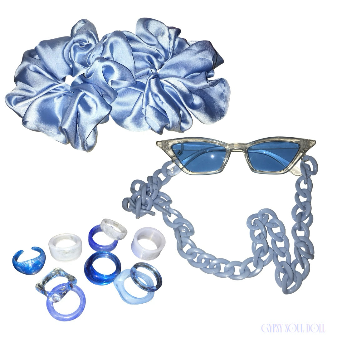 A blue hair accessory set on a white background, including two large satin scrunchies, a pair of cat-eye Ibiza fashion glasses with blue lenses and a chain lanyard strap, and several translucent blue and white retro chinky rings.