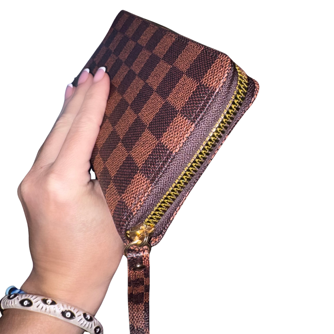 A hand holding a small zip-around wristlet clutch in brown and dark brown checkerboard monogram purse with a gold-tone zipper and wrist strap, shown against a plain white background.