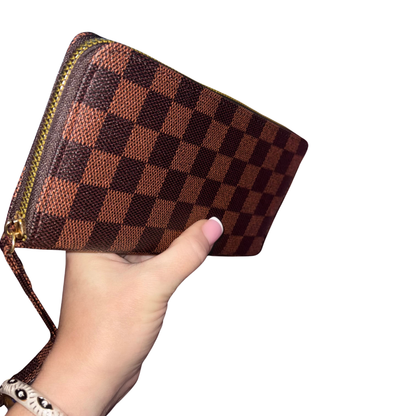 A hand holding a brown and dark brown checkerboard monogram zip-around wallet with a wrist strap, shown against a plain white background.