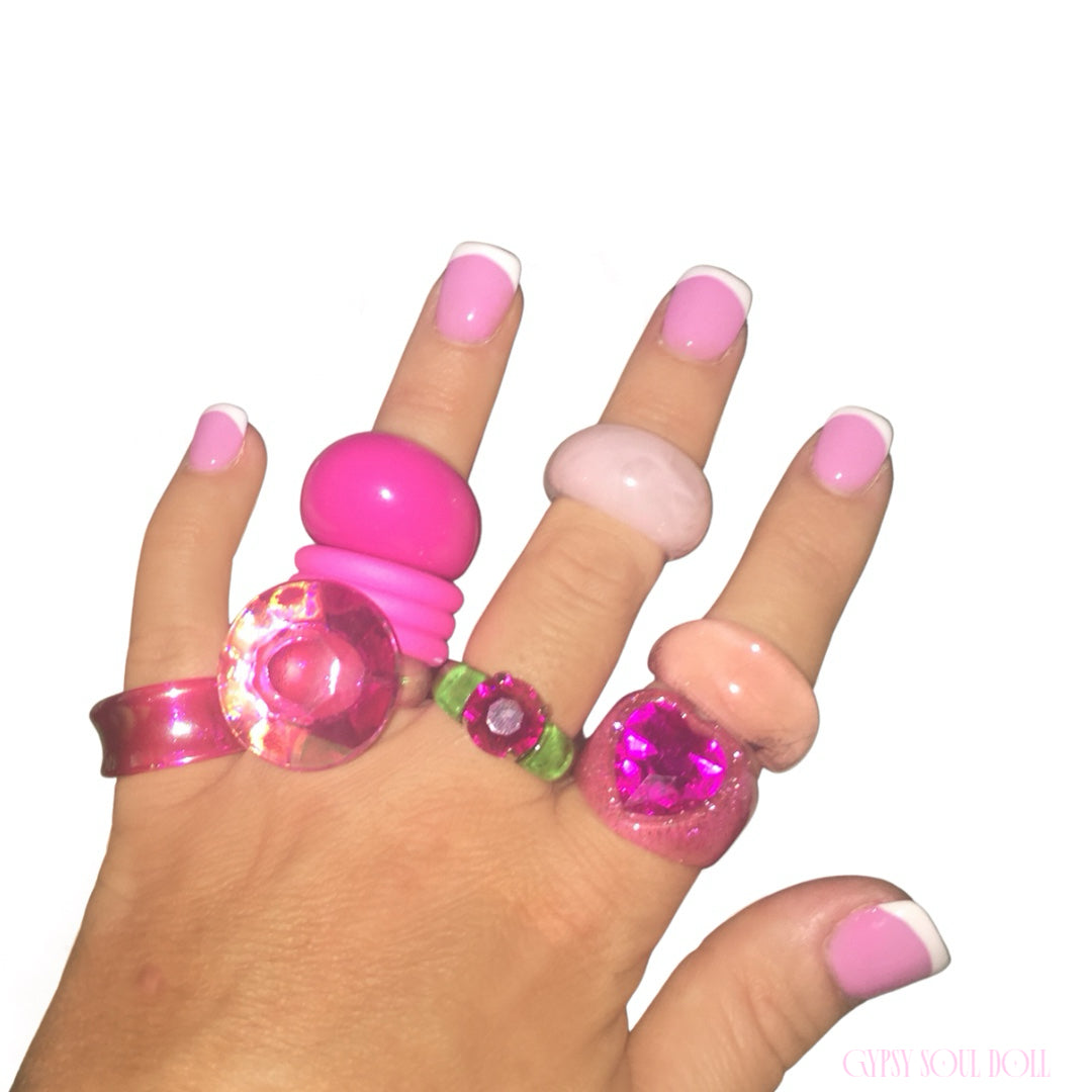 A hand with a pink and white French manicure showcasing a maximalist stack of six chunky Y2K style acrylic and resin rings in bright Barbie pink, pastel blush, transparent pink glitter, translucent magenta jewel, and clear green accents against a white background.