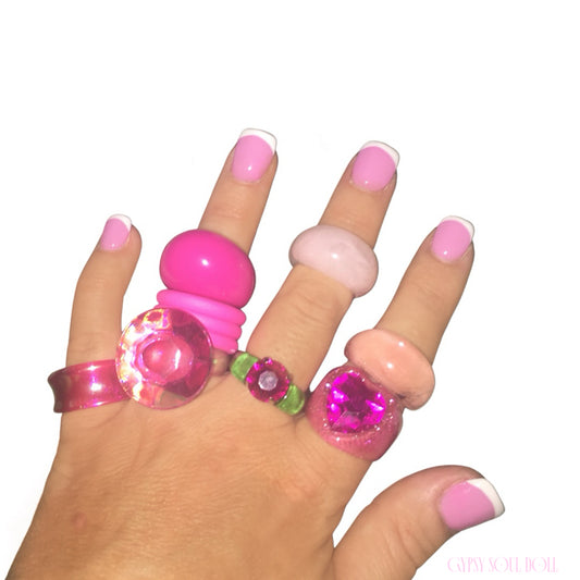 A hand with a pink and white French manicure showcasing a maximalist stack of six chunky Y2K style acrylic and resin rings in bright Barbie pink, pastel blush, transparent pink glitter, translucent magenta jewel, and clear green accents against a white background.